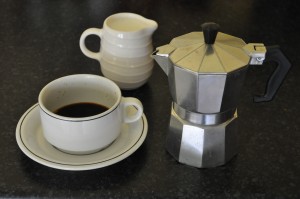 A cup of coffee, poured from a stove-top espresso machine with a pot of warm milk in the background