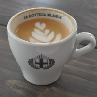A flat white from La Bottga Milanese in Leeds. The coffee is in a white, tulip cup with the words "La Bottga Milanese" written on the inside of the rim, with the cafe's logo on the front.