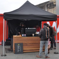 A black pop-up gazebo shelters a wooden counter holding a grinder and espresso machine