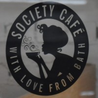 A black circle surrounding a woman in silhouette blowing on a cup of coffee. The words "Society Cafe with love from Bath" are written around the circle.
