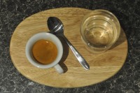 An espresso in a classic white cup, plus a glass of water, on an oval wooden platter, separated by a tea spoon.