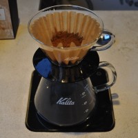 A glass Kalita Wave Filter sitting on a glass Kalita carafe which itself is on a pair of black Acaia scales. The ground coffee has been put in the filter paper, ready for brewing.