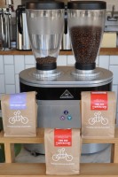 A dual-hopper Malhkonig Coffee Grinder with three bags of Tandem Coffee Roasters coffee in front of it, each with Tandem's logo of a stick-figure tandem bicycle.