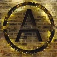 The letter A in a circle, mounted on a exposed brick wall. The circle is wrapped in fairy lights.