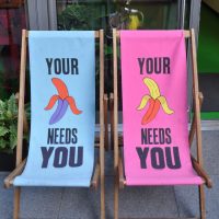 A pair of Beany Green deckchairs, blue on the left, pink on the right. Each shows a half-peeled banana with the word "YOUR" above the image, and "NEEDS YOU" underneath.