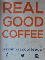The words "REAL GOOD COFFEE" in orange on white-painted brick wall. At the bottom, in blue, is Compass Coffee's social media details.