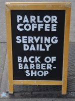 A simple blackboard with "Parlor Coffee | Serving Daily | Back of Barber-shop" written in white block capitals.