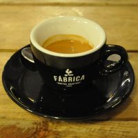 An espresso in a classic black cup, with white interior, on a black saucer. The words "Fábrica Coffee Roasters Lisboa" are written in white on the side of the cup.