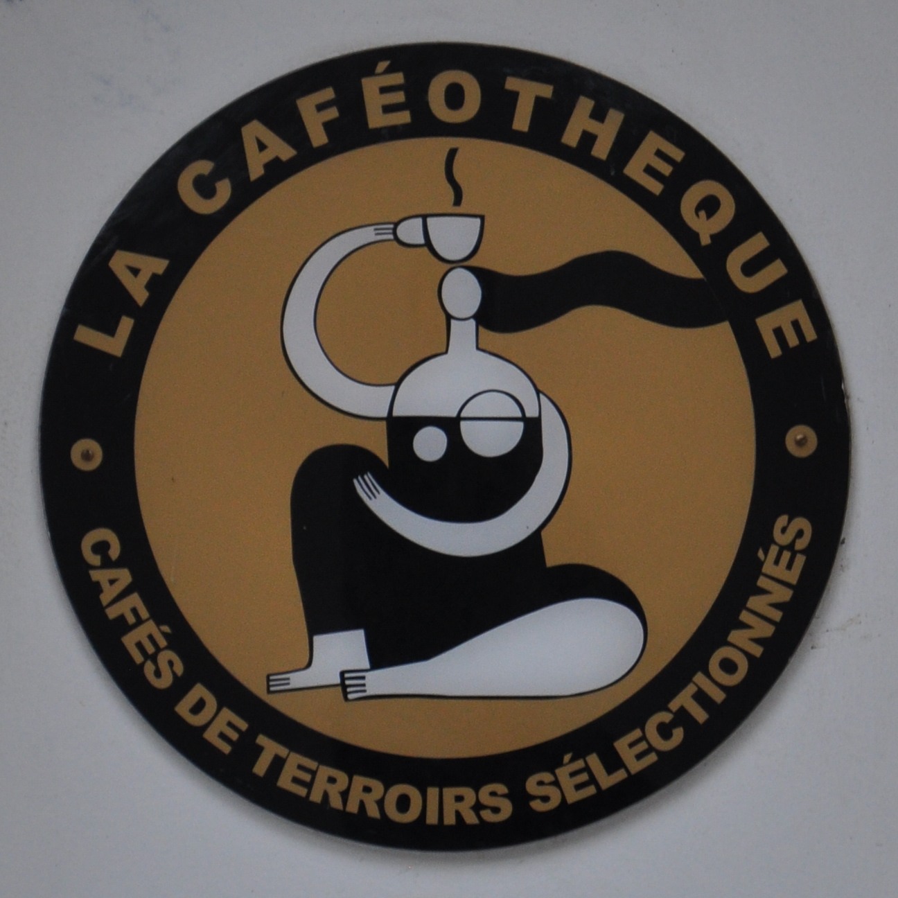 The La Caféothèque logo, a stylised picture of a woman, sitting on the floor, drawn so that her torso looks like a coffee roaster, holding an espresso cup above her head.