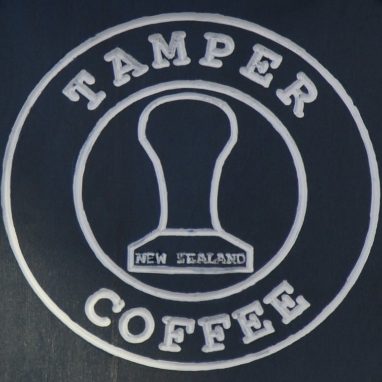 The Tamper Coffee logo, in white on black, from the sign above the door. The words "Tamper Coffee" written in a ring around the outline of tamper, with the words "New Zealand" written on its base.