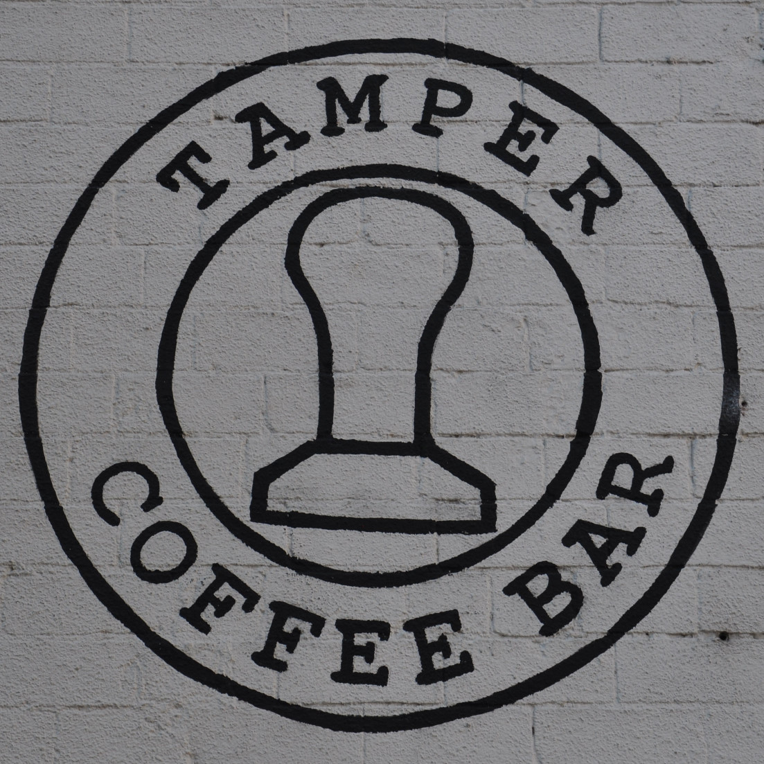 The Tamper Coffee logo painted in black on the whitewashed side of Sellers Wheel. The words "Tamper Coffee Bar" written in a ring around the outline of tamper.
