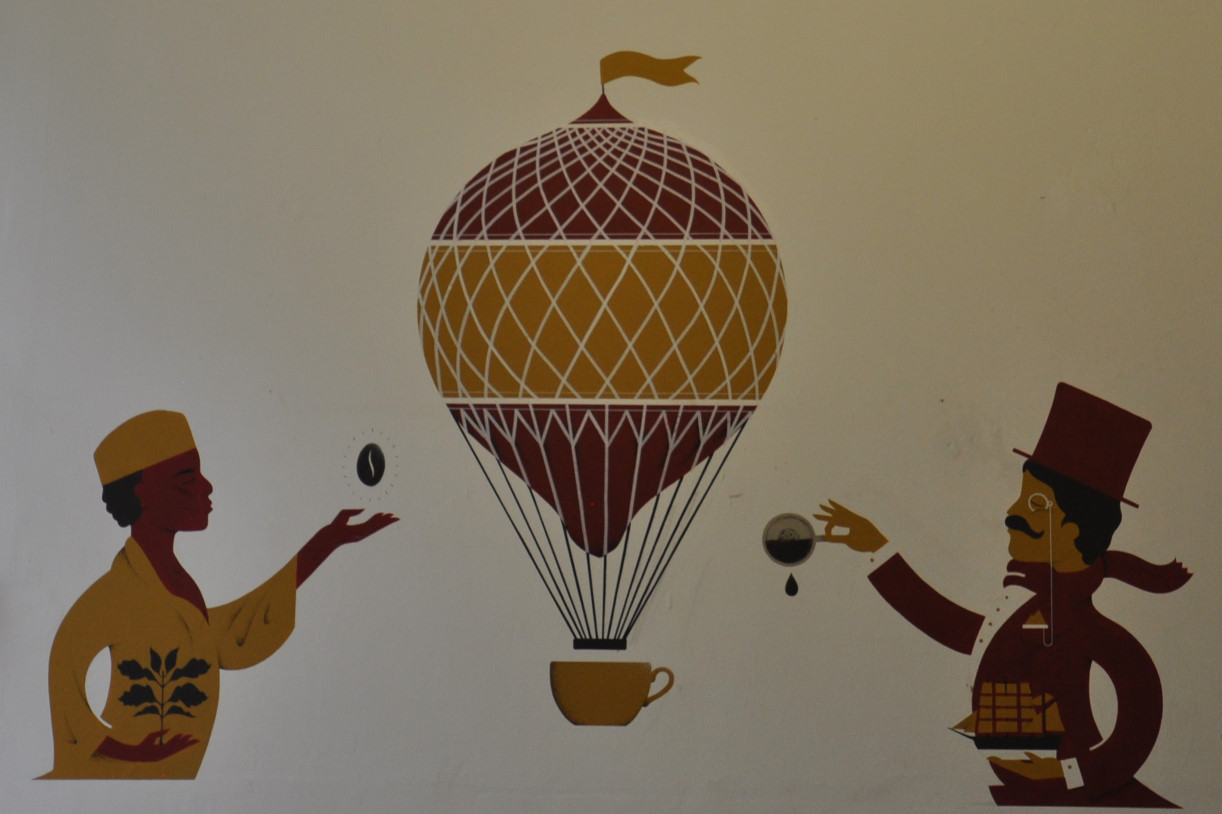 A drawing on the wall of Cafe 1901. Two figures flank an old-fashioned hot air balloon, with a coffee cup as a basket. One holds a coffee plant and coffee bean, the other, in top-hat and with monocle, holds a model sailing ship while tipping a drop of coffee out of a cup.
