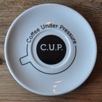 A saucer, seen above, with the outline of a cup drawn on the base of the saucer. The words "Coffee Under Pressure" are written around the circumference of the outline. In the centre is a black circle with "C.U.P." written in white in the very centre.