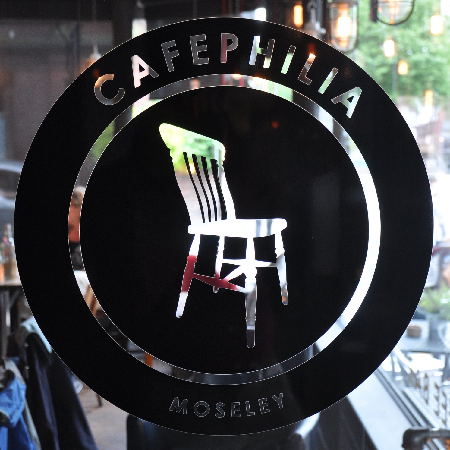 A black circle, with a clear dining room chair in the centre. "Cafephilia" is written at the top of the circle and "Moseley" is written at the bottom.