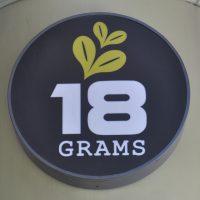 The words "18 GRAMS" in white in a black circle. Some stylised coffee beans are drawn above the 18.