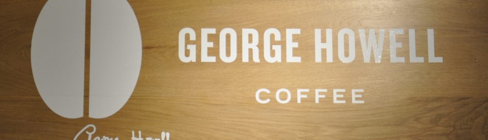 "George Howell Coffee" written in white on a wooden board, next to a silhouette of a coffee bean with George Howell's signature beneath it.