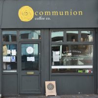The façade of Communion Coffee, with a large window on the right, the left-hand side split between the door (right) and a much smaller window (left). The yellow branding at the top stands out against the grey paintwork.