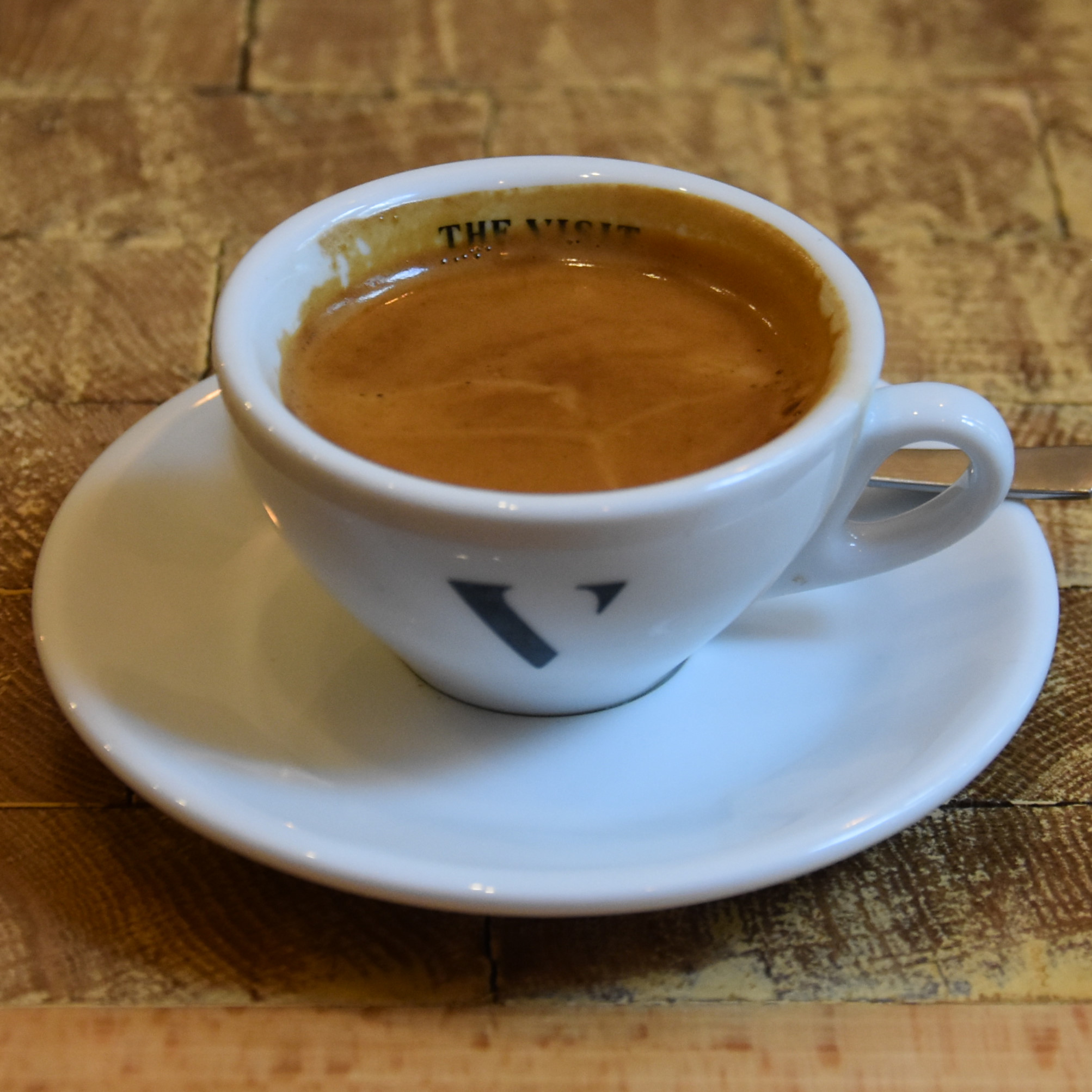 A espresso in a wide-brimmed white cup with a V on the front (for The Visit).