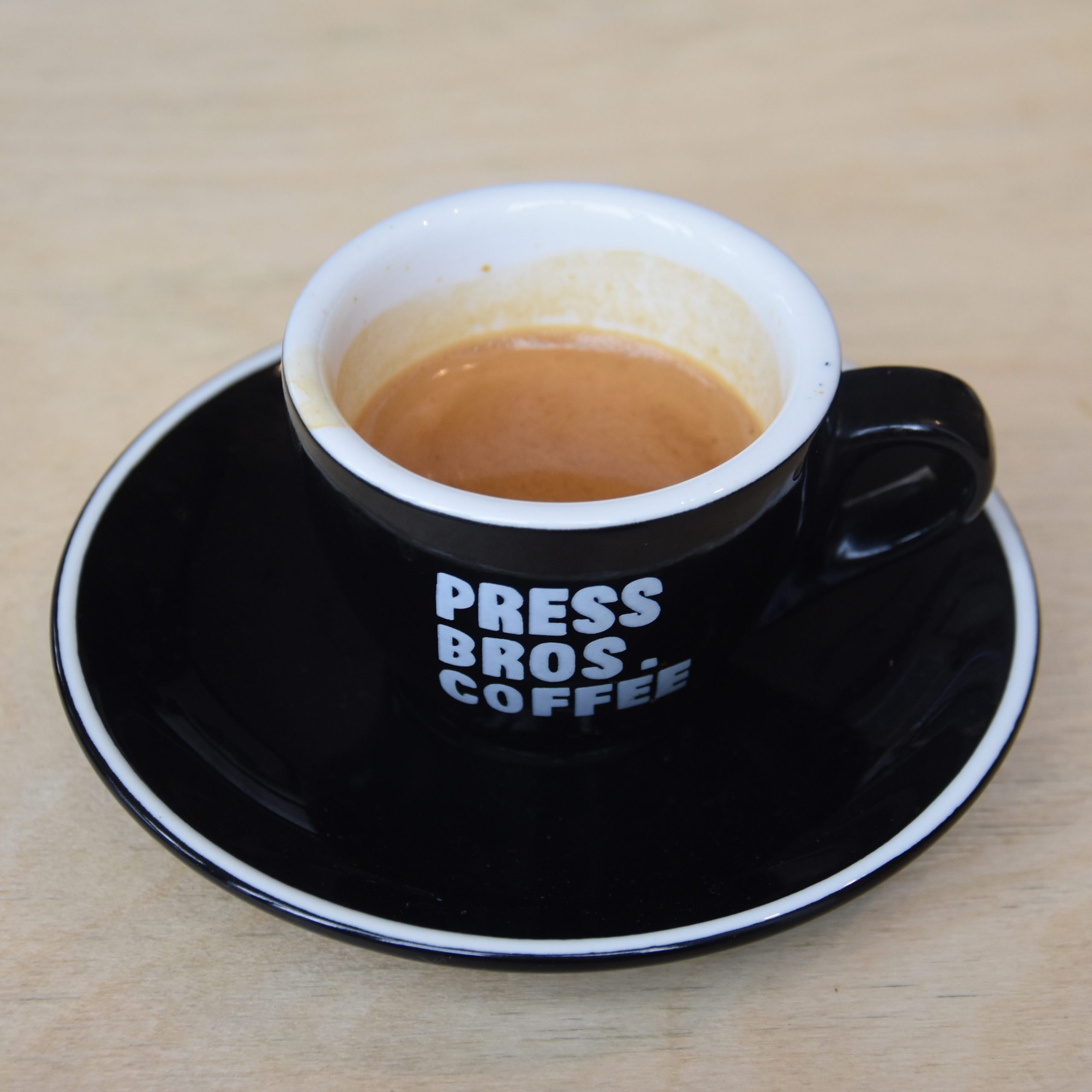 An espresso served in a classic black cup with the words "Press Bros. Coffee" on the front.