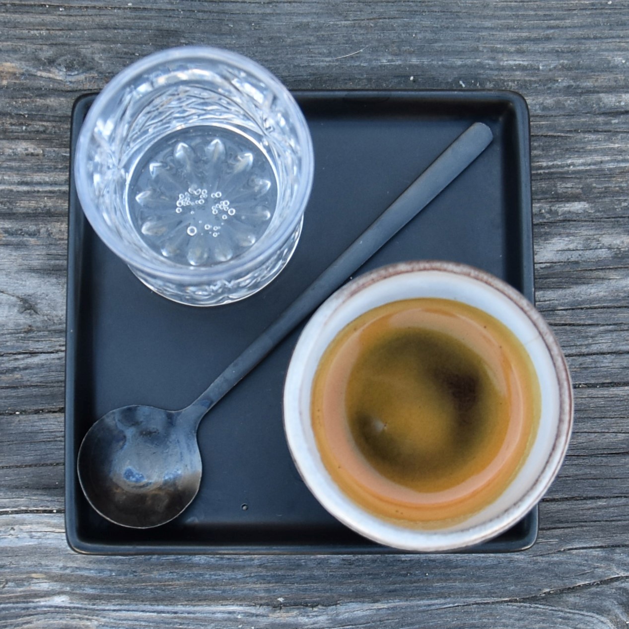 My espresso, a single-origin from East Timor, roasted by Devout Coffee and served at the coffee shop in Niles, along with a glass of water, presented on a square, metal tray, with a spoon laid diagonally between espresso and glass.