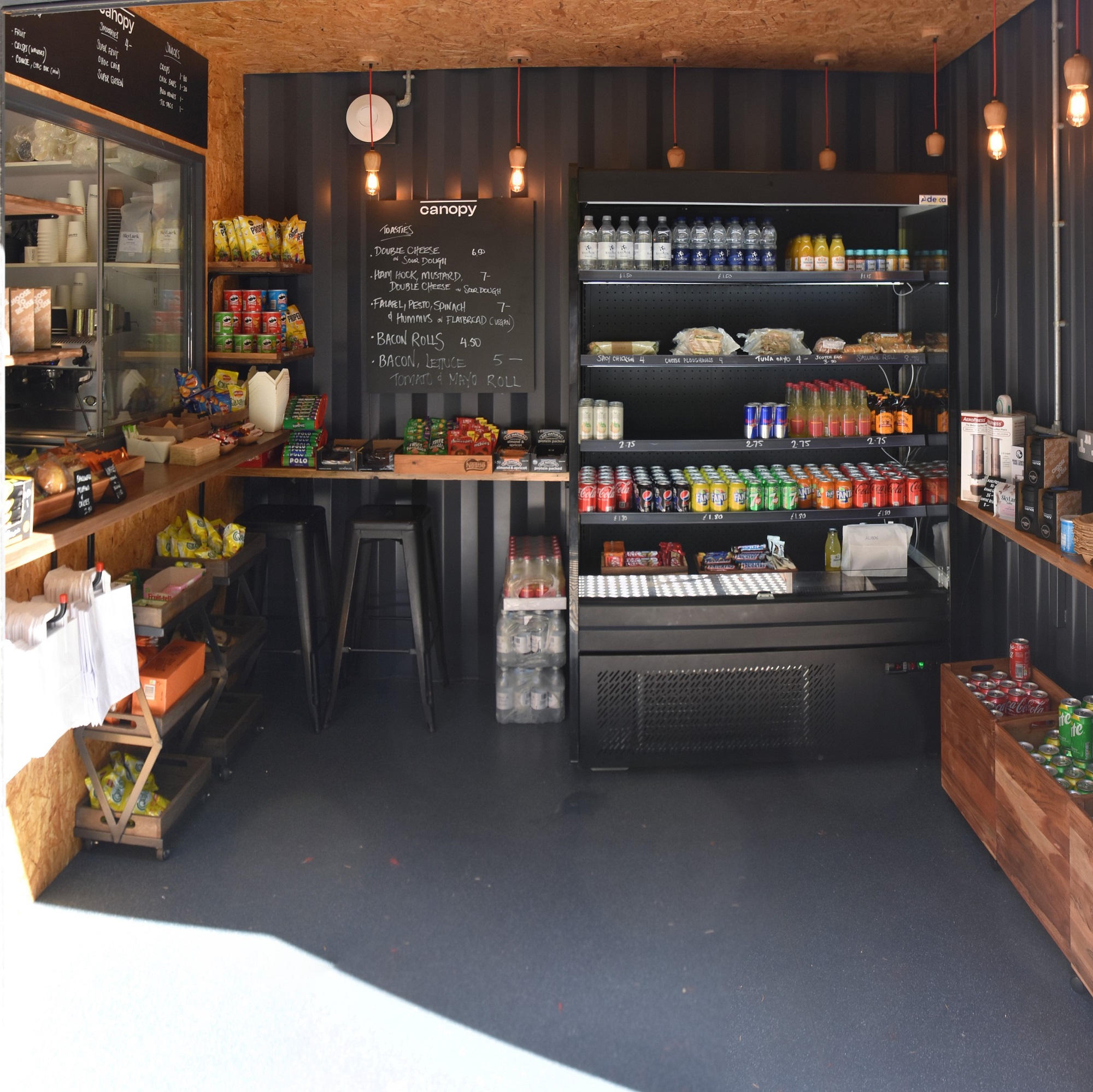 The interior of Canopy Coffee, London Square, a cosy coffee cabin/container, with the counter on the left and shelves full of goodies lining all three walls.