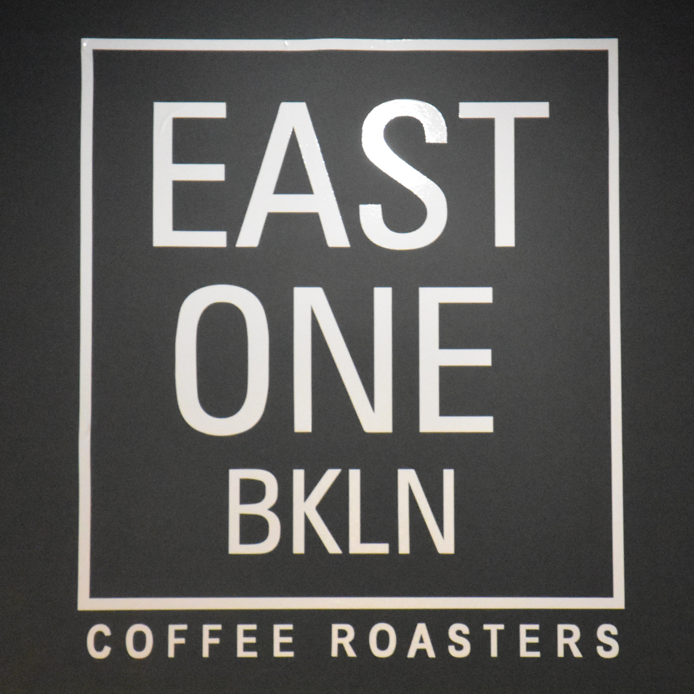 Detail from the wall of East One Coffee Roasters in midtown Manhattan: "EAST ONE BKLN" in white on black, outlined with a white square, with "COFFEE ROASTERS" underneath.