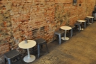 The seating is a row of two-person round tables against the bare-brick wall on the left.